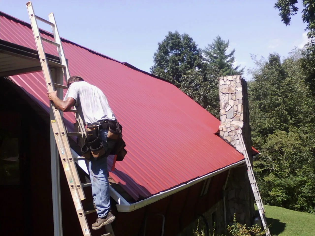 Roofer on a ladder beside a red metal roof and stone chimney — Soffit & Fascia Repair by Roofing Brothers in Drexel